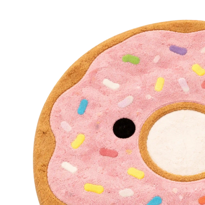 A close-up of a donut-shaped plush or cushion with pink frosting, multicolored sprinkles, a brown edge, and a white center hole on a white background.