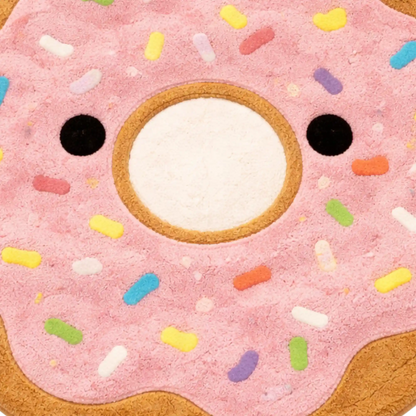 A close-up of a donut-shaped rug or mat with pink frosting, multicolored sprinkles, and a white center with black circular eyes.